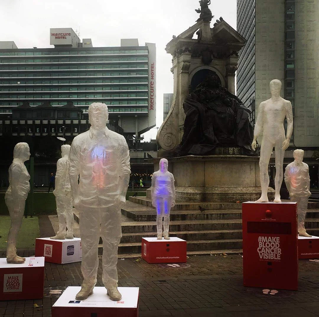 Grupa półprzezroczystych postaci na postumentach obok pomnika w kampanii Make Blood Cancer Visible.