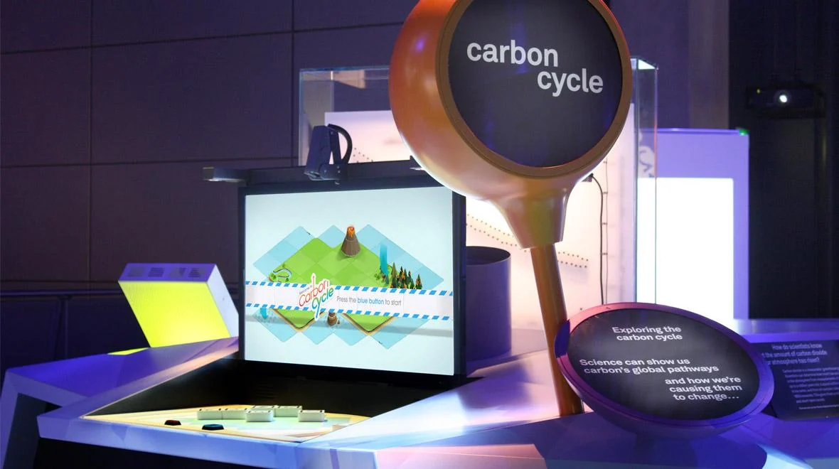 Eksponat Carbon Cycle z ekranem, kontrolerami na stole i okrągłym pomarańczowym znakiem.
