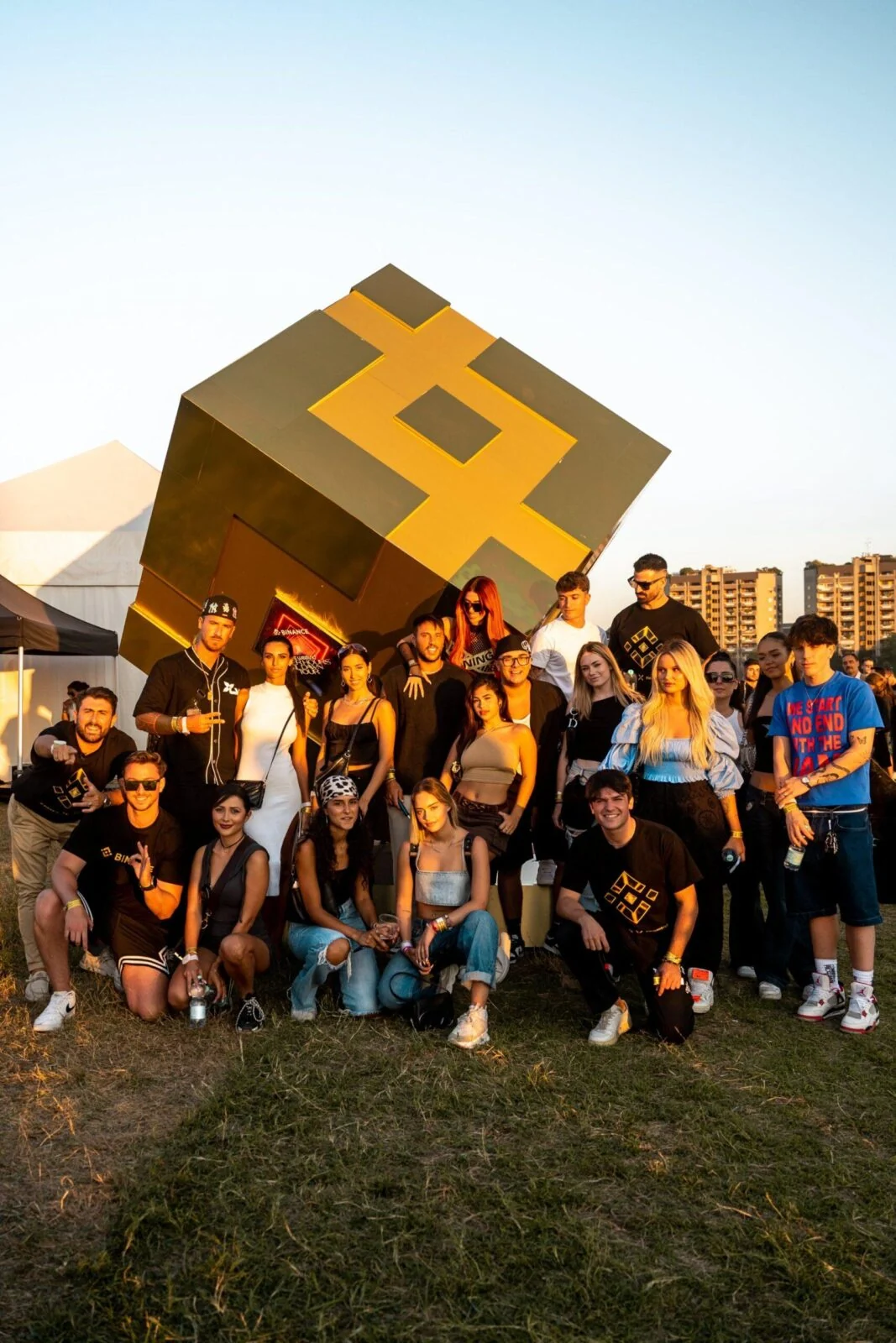 Grupa pozuje przed dużym złotym sześcianem Binance podczas wydarzenia plenerowego.