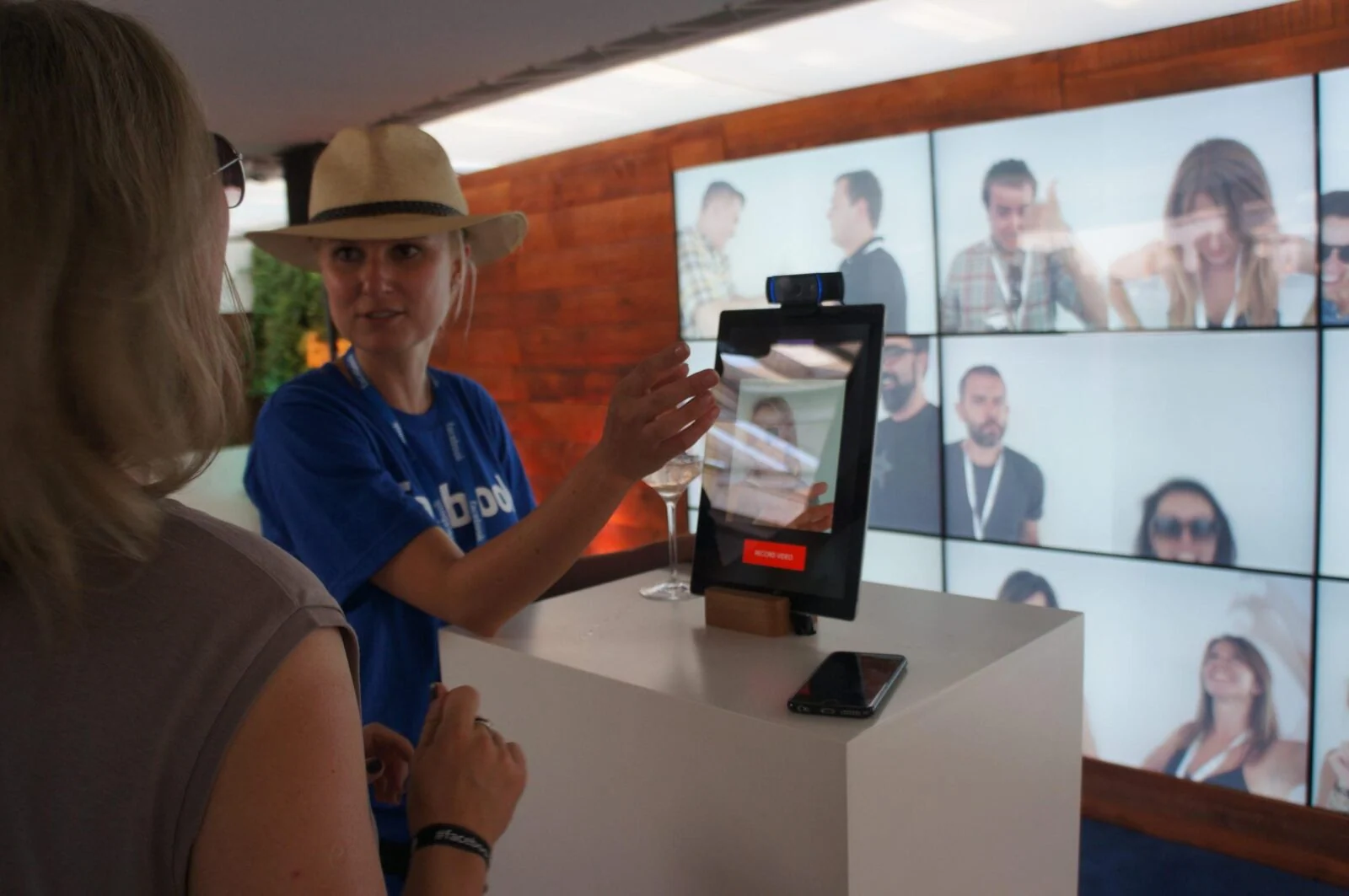 Zwiedzający i pracownik korzystają z kiosku z kamerą przed ścianą portretów wideo w strefie Facebook.
