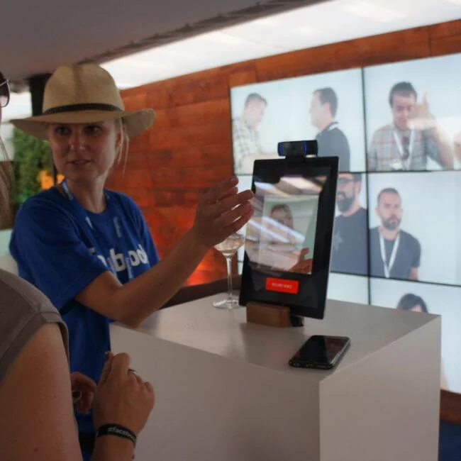 Zwiedzający i pracownik korzystają z kiosku z kamerą przed ścianą portretów wideo w strefie Facebook.