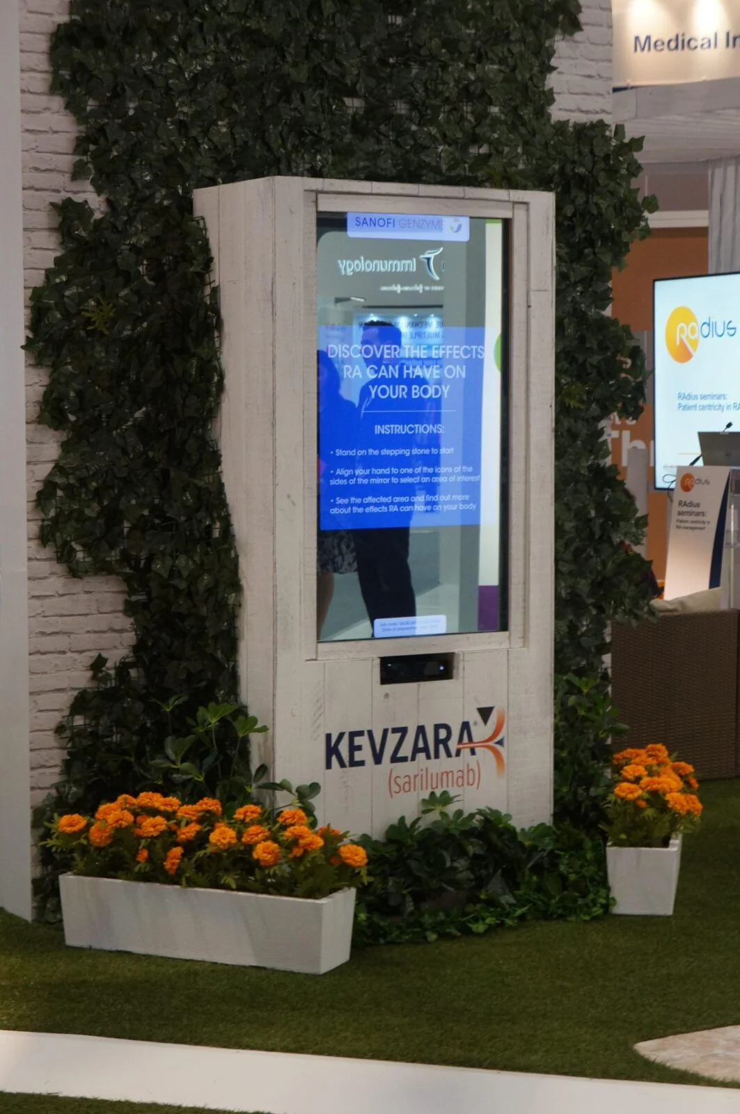 Kiosk z interaktywnym lustrem rentgenowskim w zielonej ścianie roślin na stoisku wystawowym.