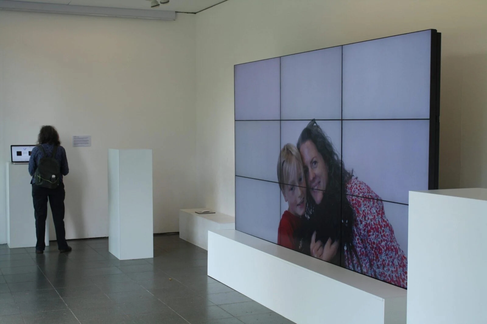 Instalacja galeryjna z dużą ścianą wideo pokazującą uśmiechniętych uczestników.